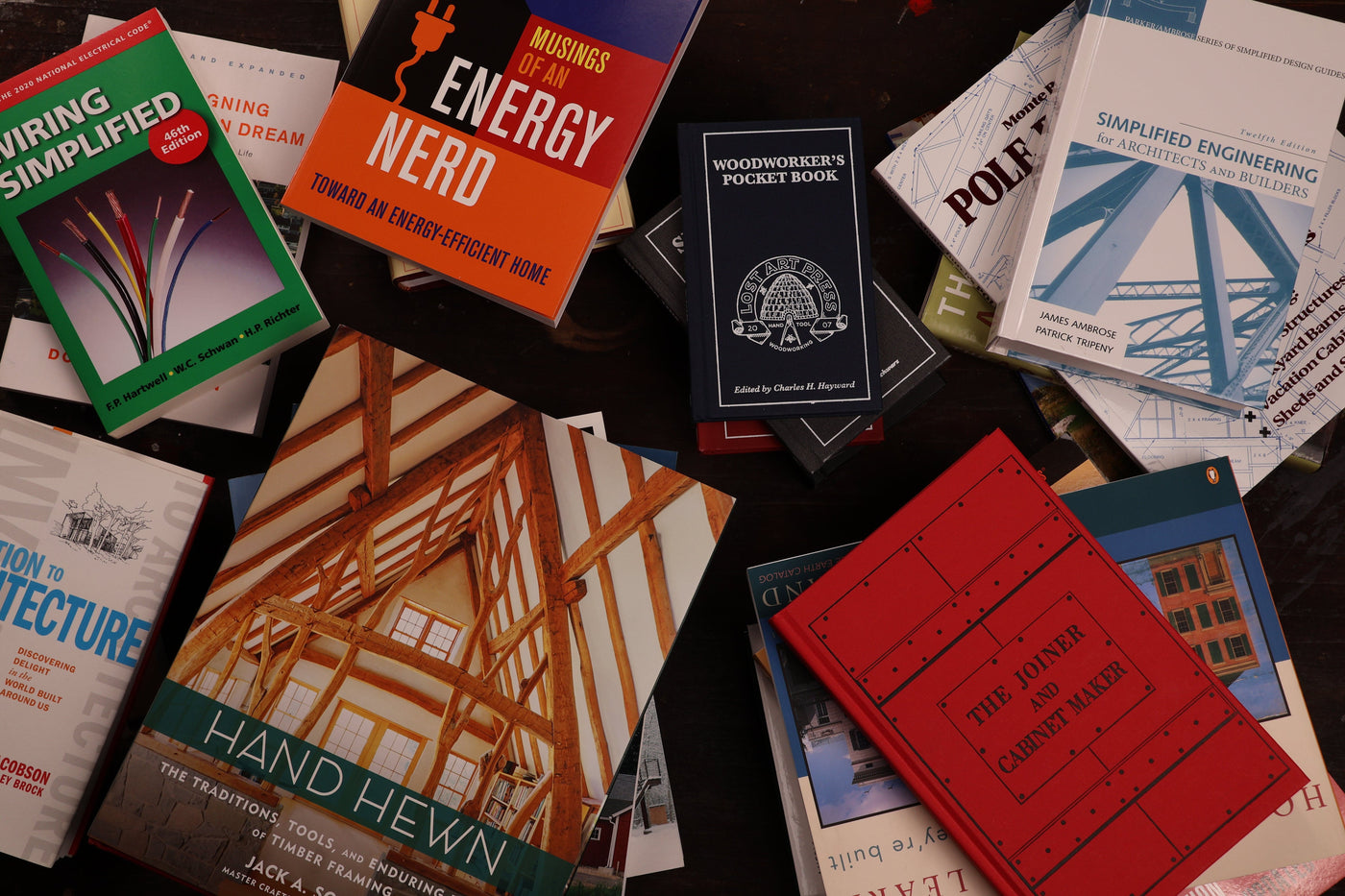 A collection of engineering, architecture, and woodworking books spread out on a dark surface, including titles like Hand Hewn and Musings of an Energy Nerd—plus guides on timber framing tools from the Shelter Institute and shelter tools.