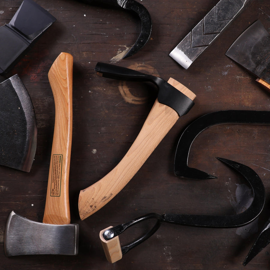 A variety of woodworking and blacksmithing tools, including timber framing tools and axes with wooden and metal handles, are arranged on a dark, worn wooden surface reminiscent of a shelter institute workshop.