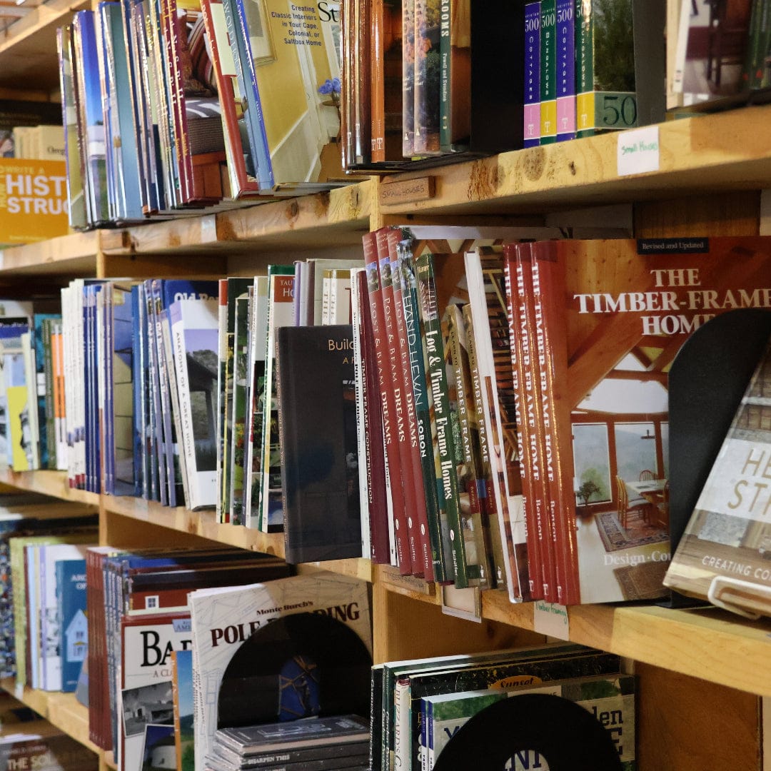 Books are neatly arranged on wooden shelves in a bookstore or library. Most books are standing upright, showcasing colorful covers and spines, with titles about architecture, home building, and timber framing tools visible.