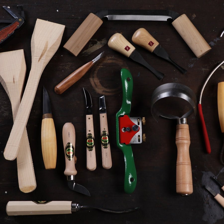 Various wood carving tools and unfinished wooden spatulas are arranged on a dark wooden surface. These quality shelter tools include knives, gouges, a spokeshave, and a draw knife, all with sturdy wooden handles.
