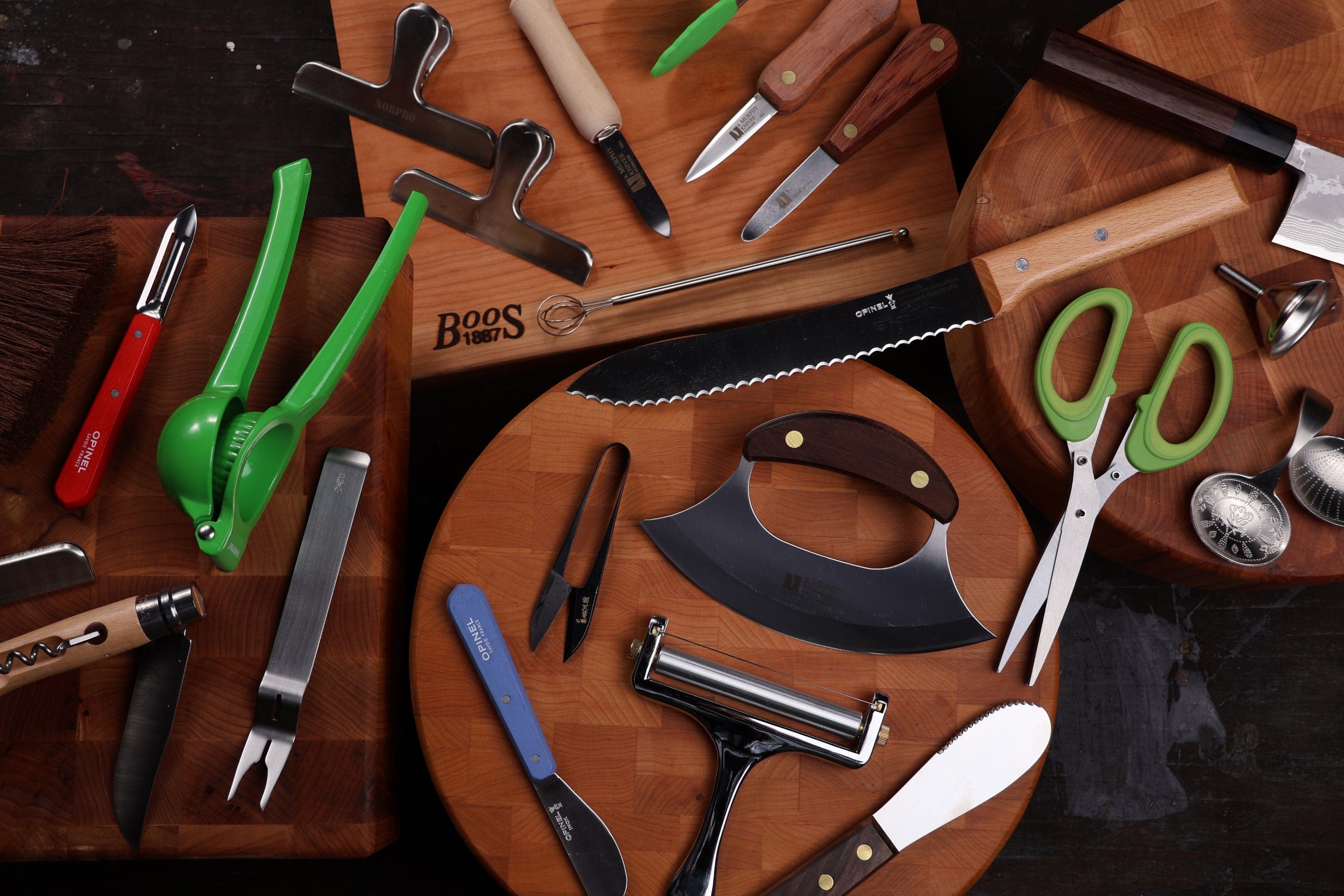 A variety of kitchen tools, including knives, scissors, peelers, a citrus squeezer, and cutting boards—reminiscent of shelter tools—are arranged neatly on a dark surface. The utensils have colorful handles and metallic blades.