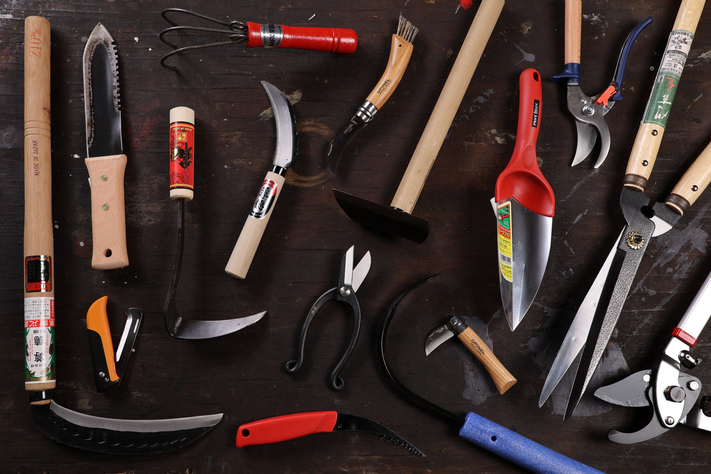 A variety of gardening and timber framing tools, including pruners, sickles, hand saws, shears, and hoes, are arranged on a dark wooden surface. Some are classic shelter tools from Shelter Institute, with handles in wood, plastic, and metal.