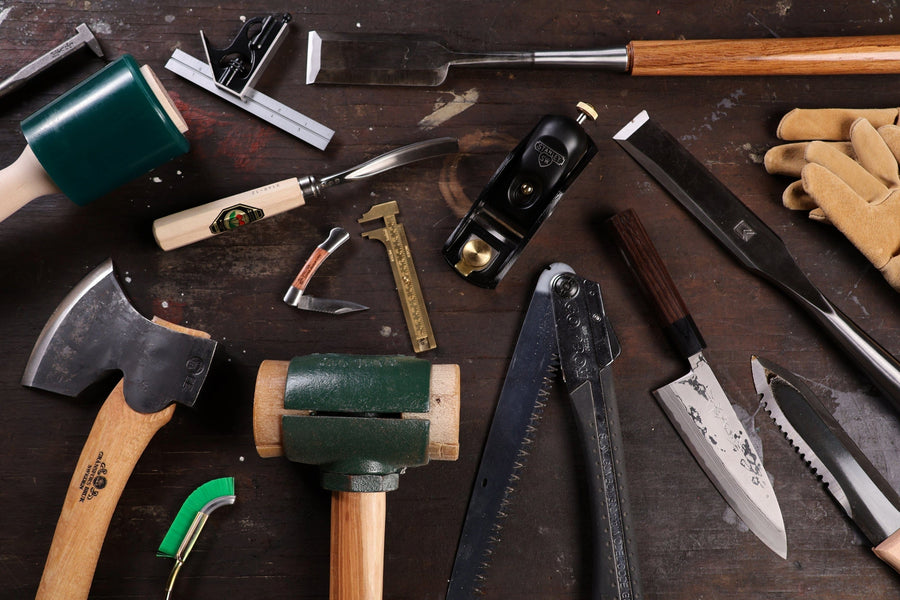 A variety of woodworking tools, including timber framing tools like hammers, axes, saws, knives, rulers, gloves, and chisels, are arranged on a dark wooden workbench, inspired by shelter institute craftsmanship.
