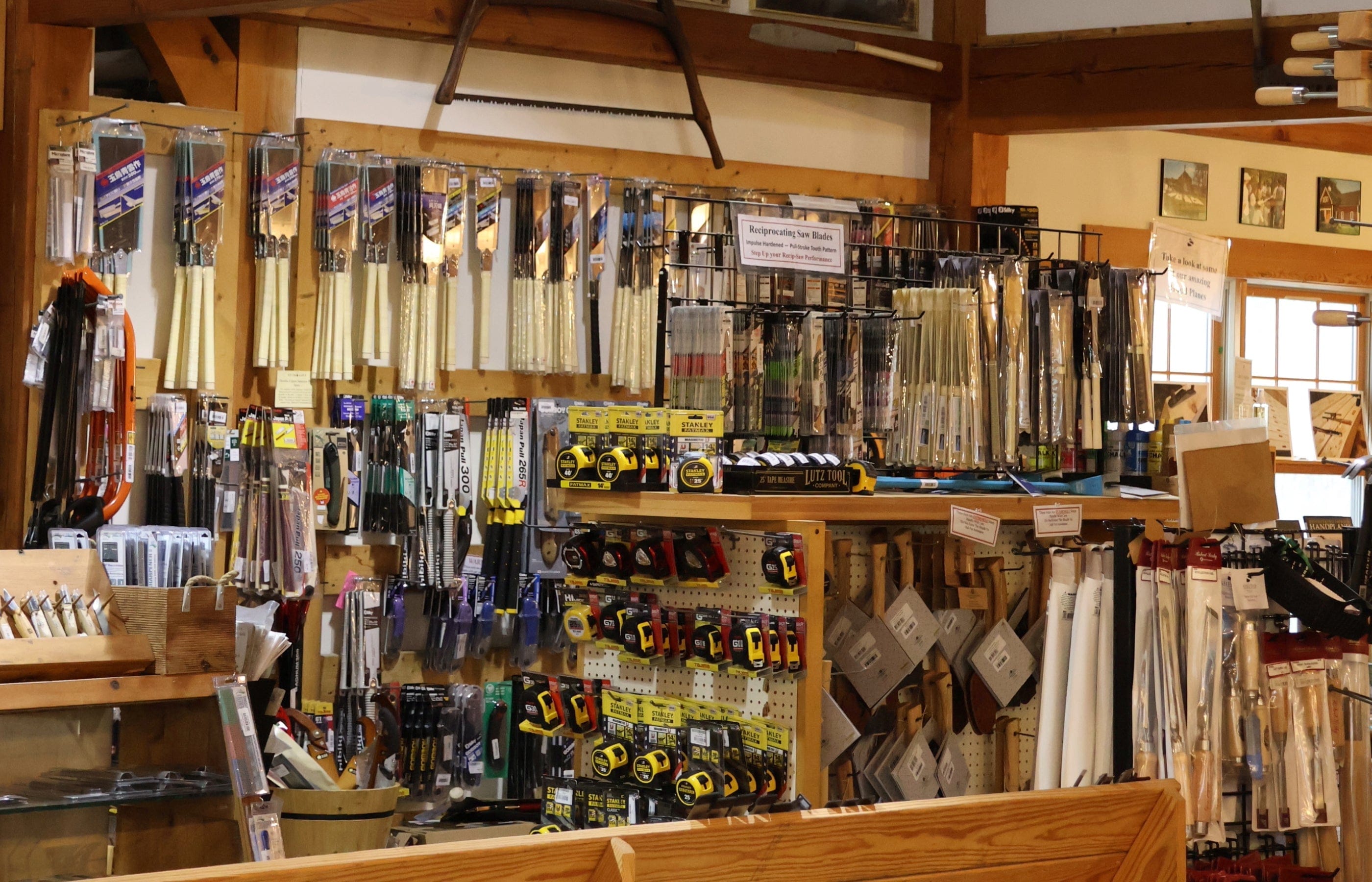 A hardware store display with various hand tools, including timber framing tools, tape measures, saw blades, files, and paintbrushes, organized on racks and pegboards against a wooden wall.
