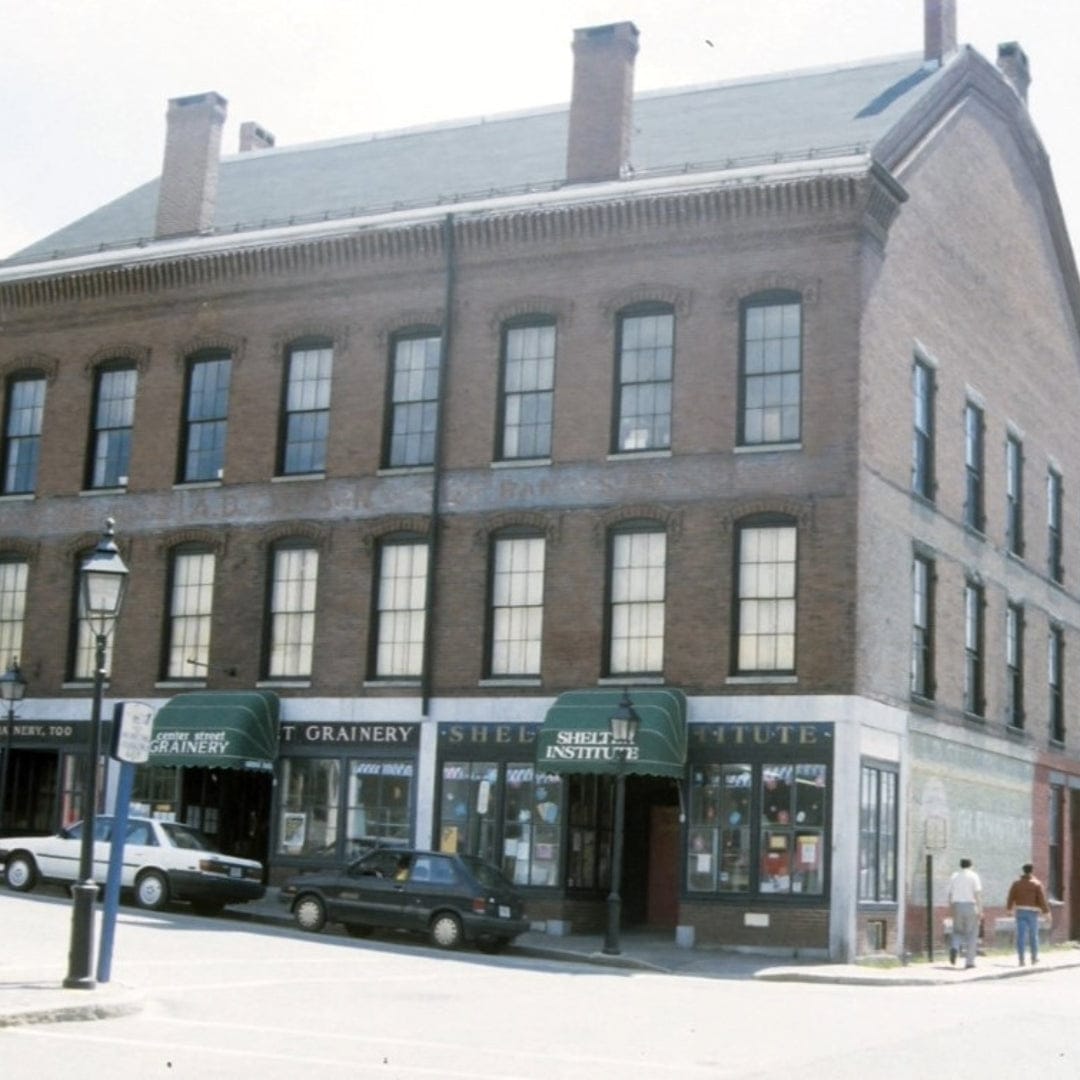 A large brick building on a street corner with tall windows and green awnings reading “SHELBURNE INSTITUTE” and “SALT GRANERY,” home to shelter tools and timber framing tools. Several parked cars and a streetlamp are visible in front.