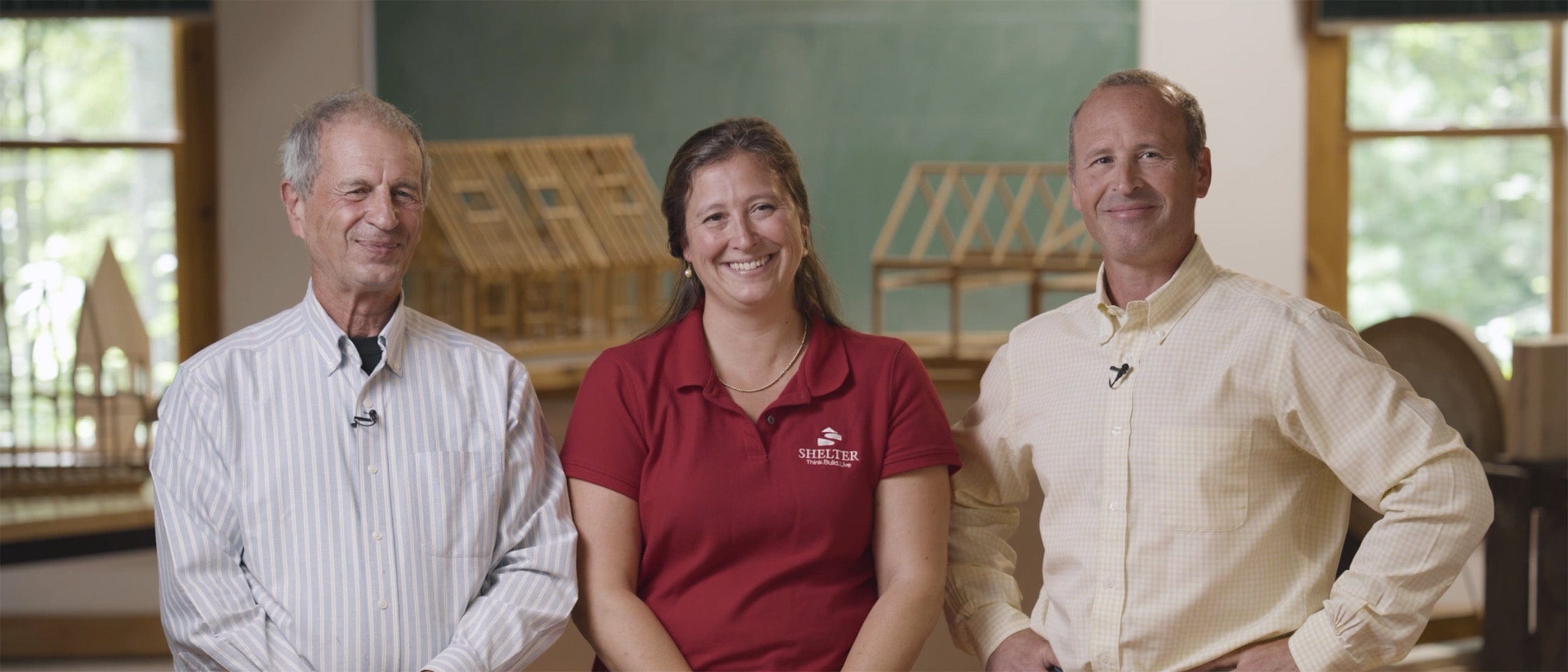 Three people stand indoors, smiling at the camera. The group is gathered at Shelter Institute, with timber framing tools and wooden model structures visible in the background.