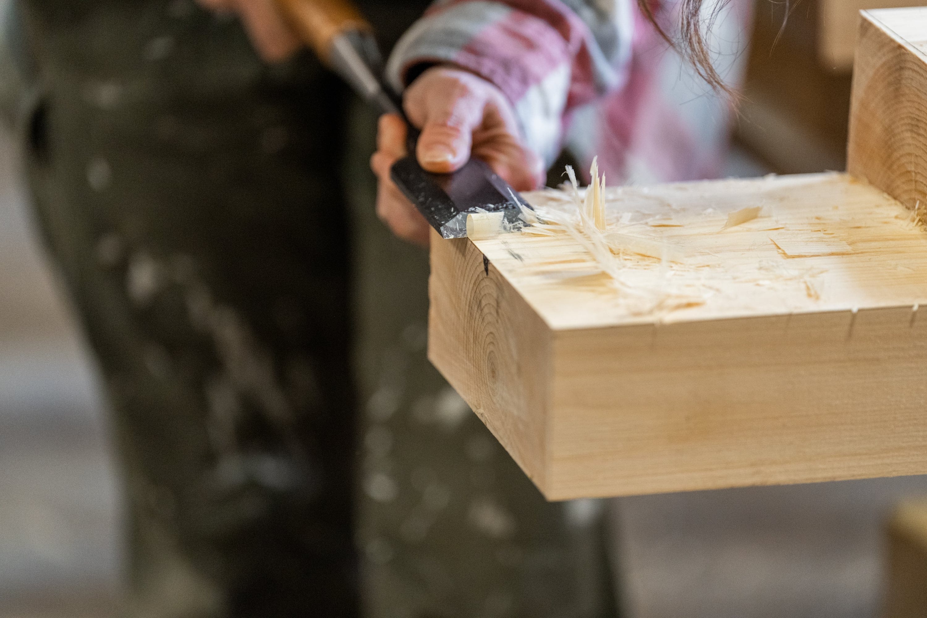 A person uses a chisel to carve and smooth the edge of a piece of light-colored wood, with wood shavings visible. The person is wearing a plaid shirt and dark pants.