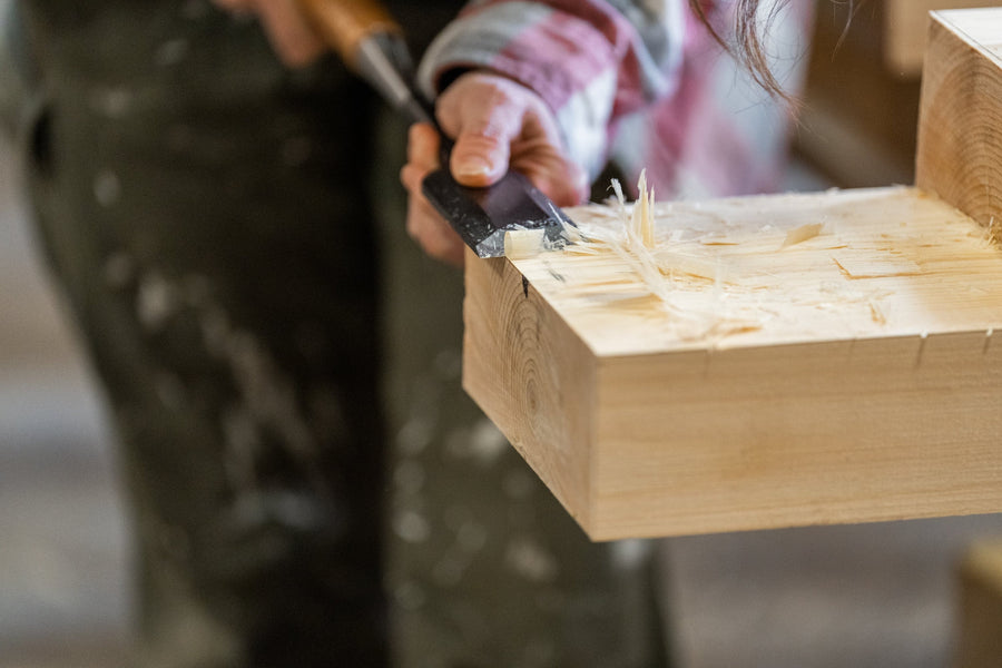 A person uses a chisel to carve and smooth the edge of a piece of light-colored wood, with wood shavings visible. The person is wearing a plaid shirt and dark pants.