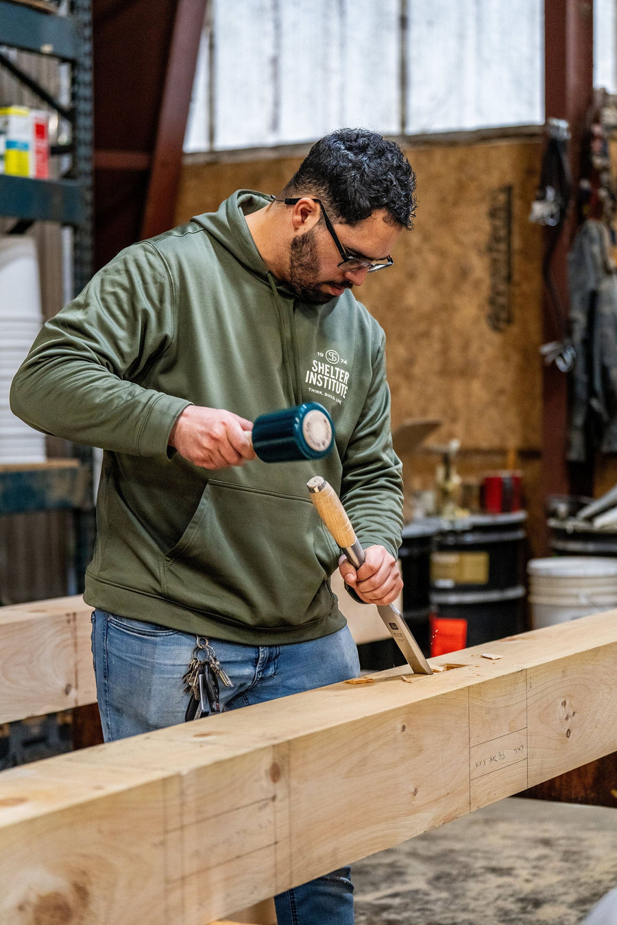 A man wearing glasses and a green hoodie uses a mallet and chisel to work on a large wooden beam in a workshop. Tools and materials are visible in the background.