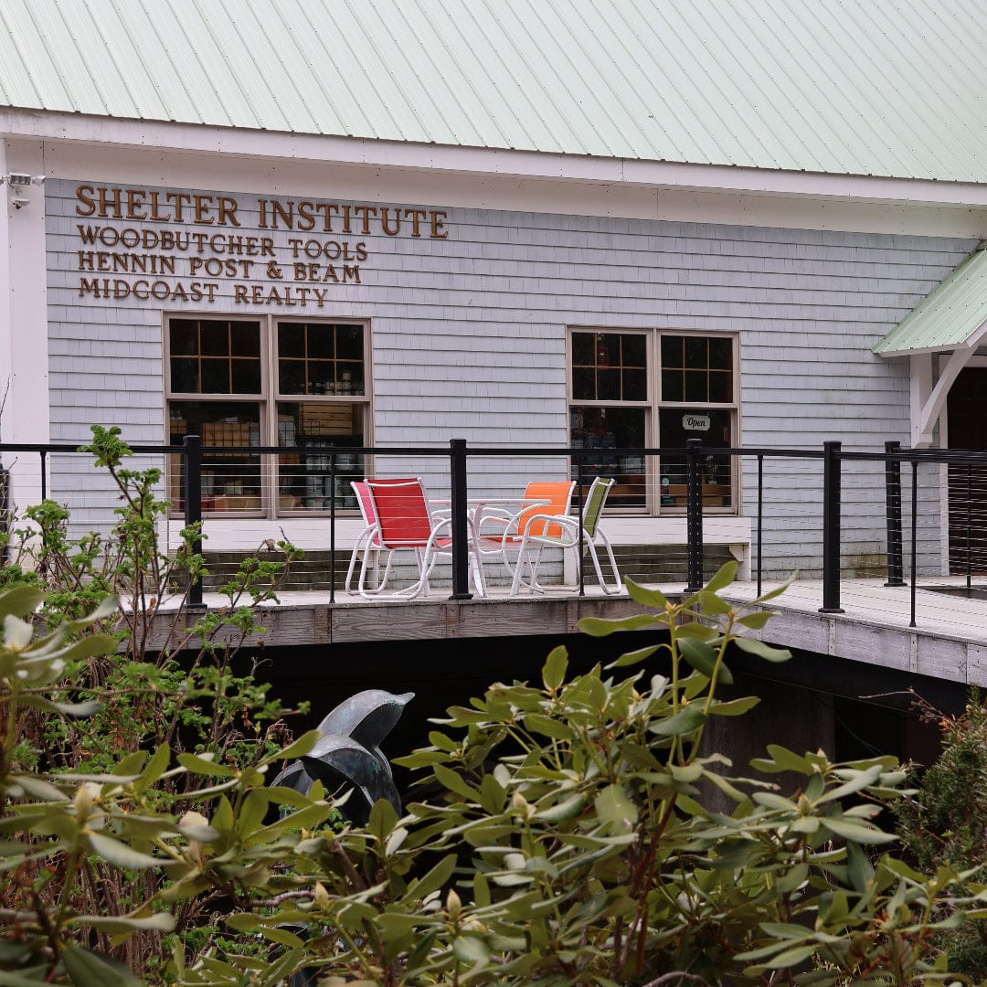 A gray building with a green roof features a sign for Shelter Institute, known for shelter tools and timber framing tools. Three colorful chairs sit on a deck in front, with plants and bushes in the foreground.