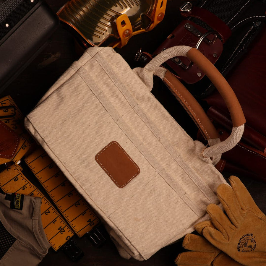 A cream-colored canvas tool bag with brown leather accents sits among various shelter tools, including gloves, a measuring tape, a hard hat, and belts, on a dark surface.