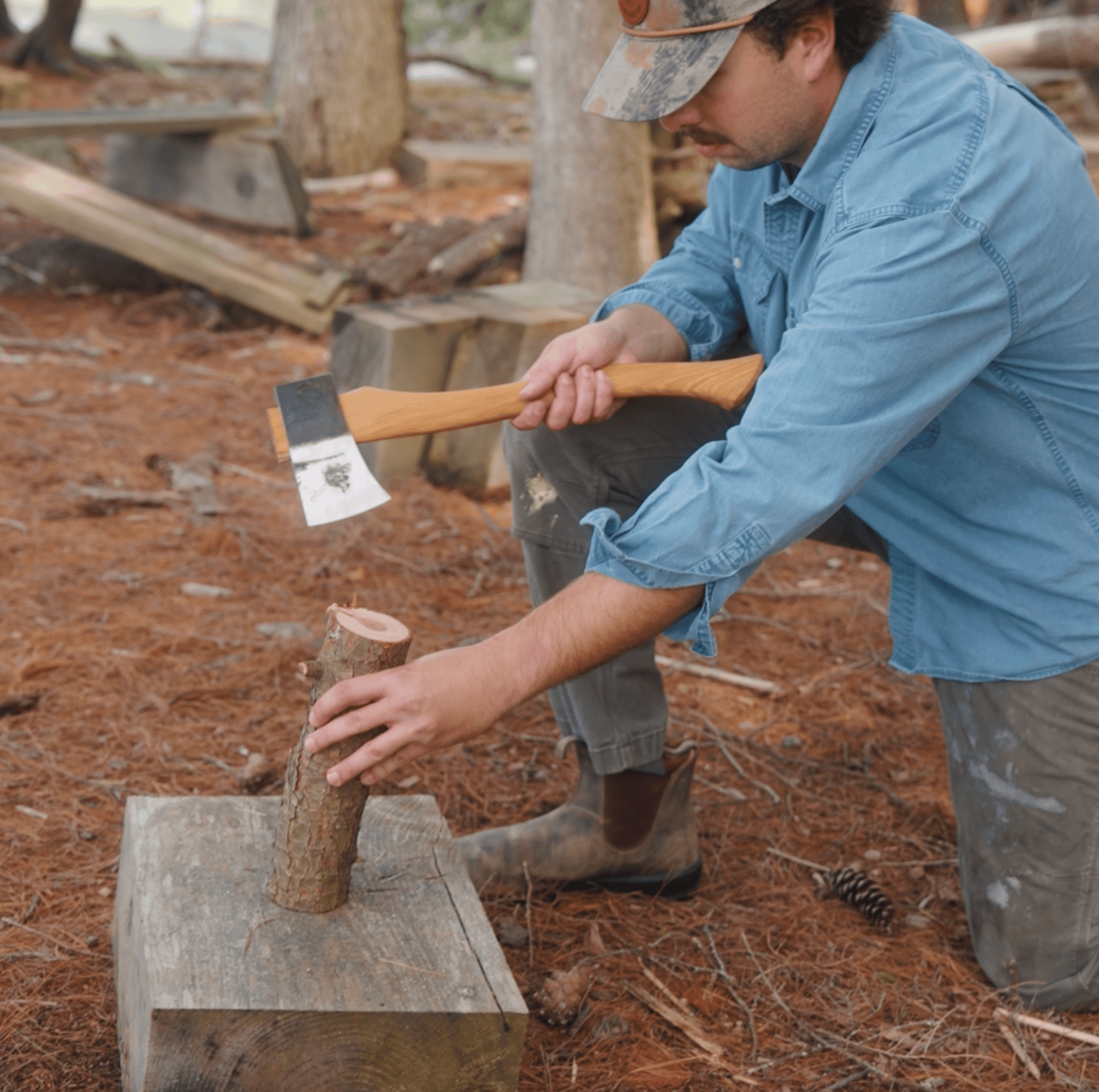 A person in a blue shirt and cap kneels outdoors, holding an axe above a small log placed on a tree stump, preparing to chop wood in a forested area.