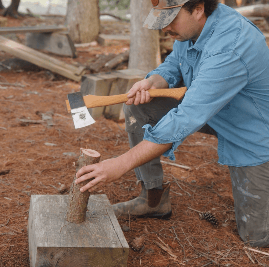 A person in a blue shirt and cap kneels outdoors, holding an axe above a small log placed on a tree stump, preparing to chop wood in a forested area.