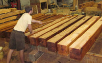 In a workshop filled with tools and materials, a man precisely measures large wooden beams to ensure accuracy for improved water resistance, using HNF's Heritage Natural Finishes Oil Select Interior & Exterior.