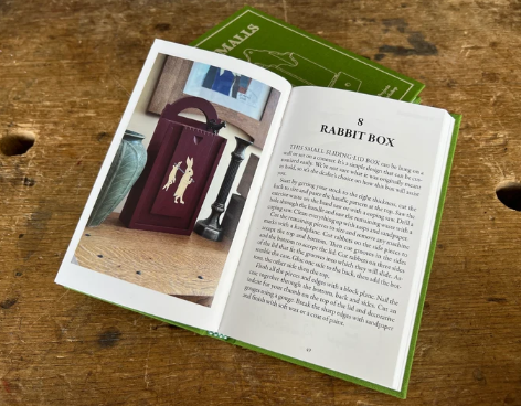 An open book titled “Smalls: 18 Well-designed, Easy Projects for an Afternoon in the Workshop” by Shelter Institute rests on a wooden surface, displaying a page featuring a maroon rabbit-adorned box project with instructions reminiscent of classic woodworking guides. A green pocketbook peeks out from underneath.