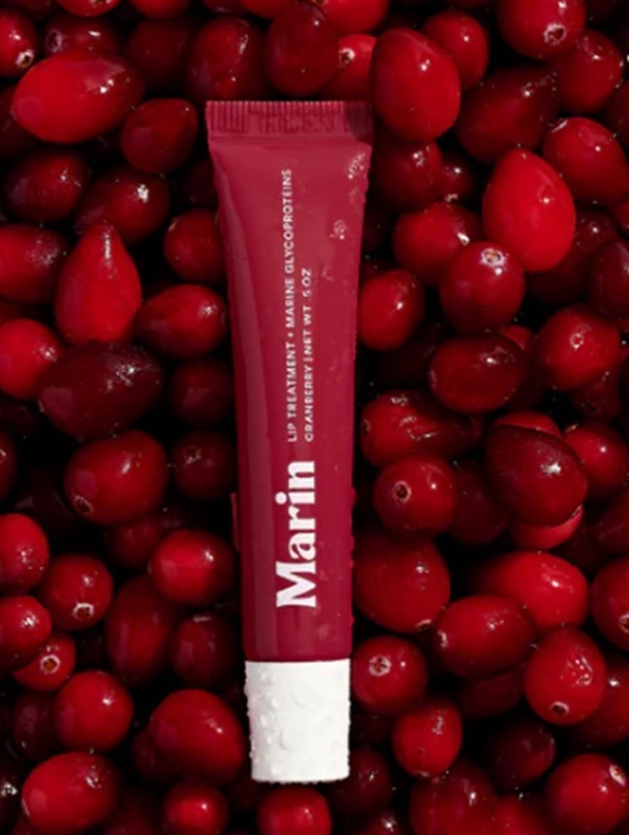 A red tube of Marin Lip Treatment by Marin, infused with cranberry fruit extract and Marine Glycoproteins, rests on fresh cranberries, blending with the vibrant red berries in the background.