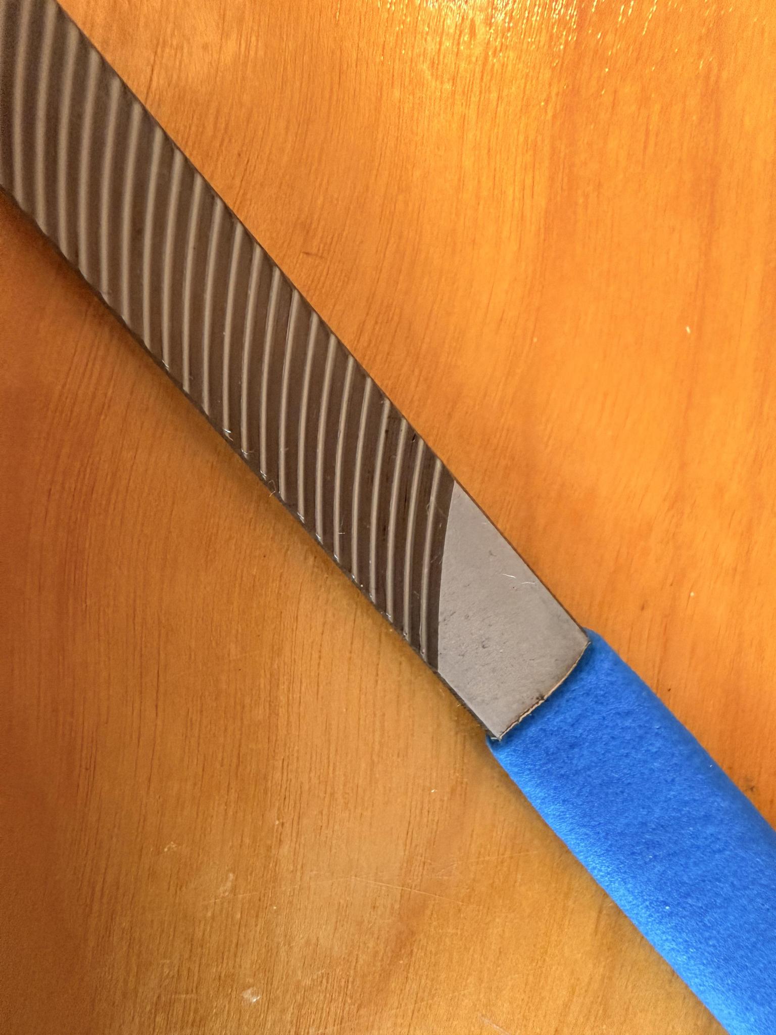 A close-up of the Iwasaki Draw File by Iwasaki, featuring diagonal grooves and a blue handle on wood—a top choice for wood carving and achieving a smooth surface finish.