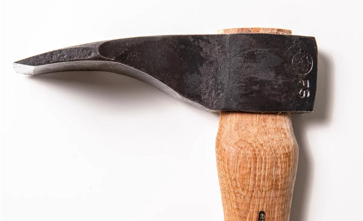 A close-up photo of the Shelter Institute Small Straight Adze 56mm, featuring a black metal head and a wooden handle, displayed against a white background, evokes the craftsmanship found in traditional log building and toolmaking.