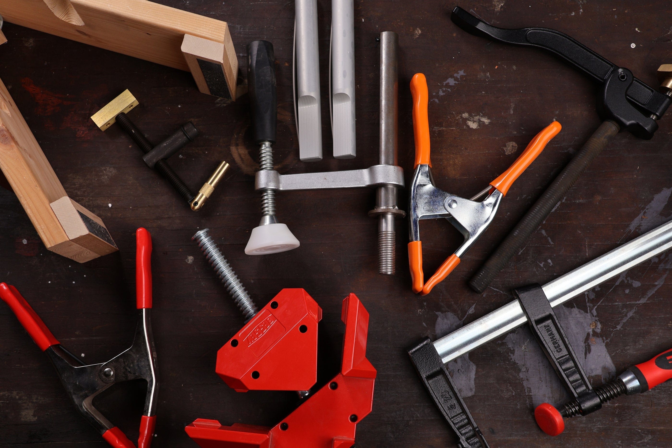 Various woodworking clamps and tools, including C-clamps, quick-grip clamps, and timber framing tools from Shelter Institute, are scattered on a dark, worn workbench surface.