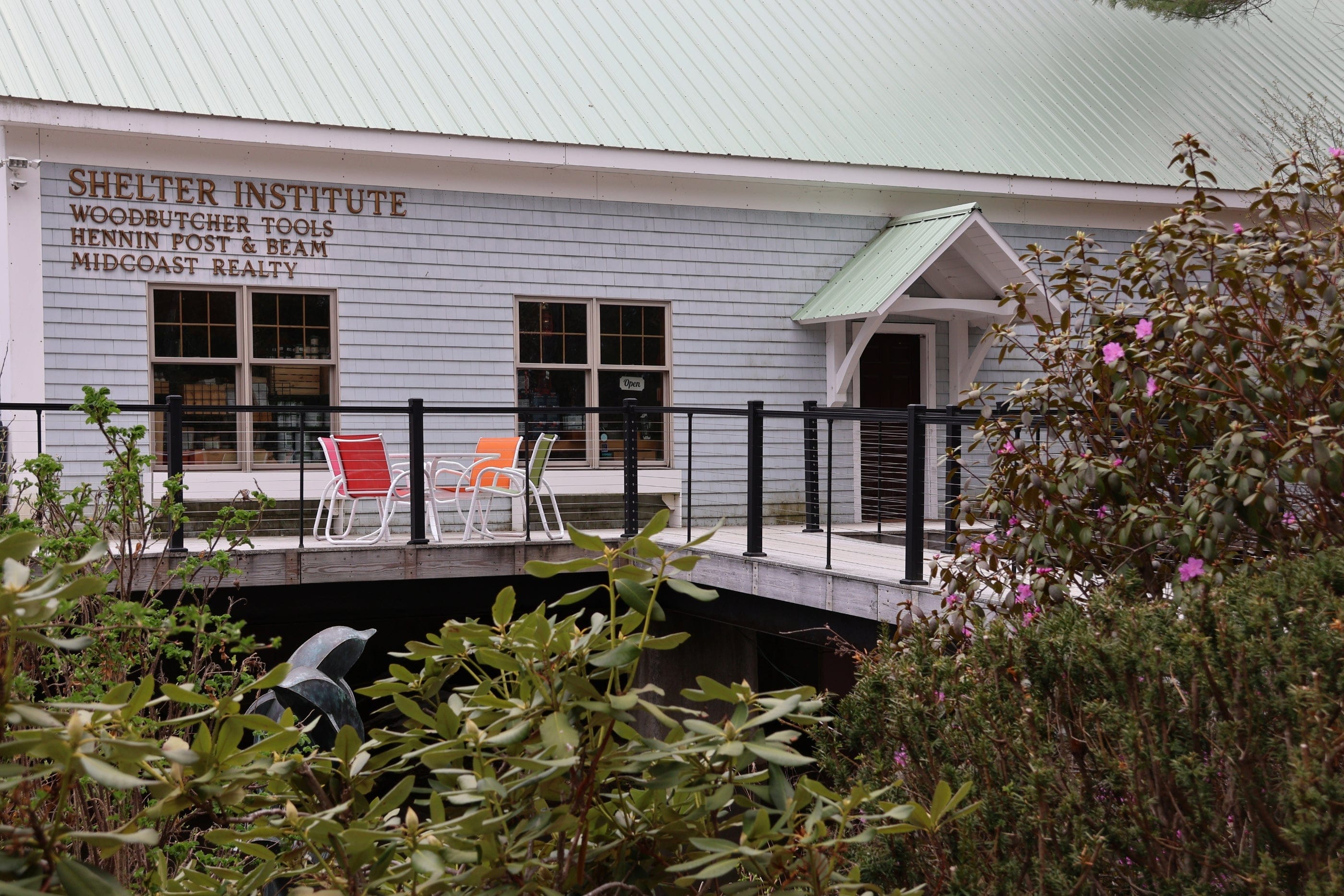 A gray building with a green roof features signs for Shelter Institute, known for timber framing tools, Woodbutcher Tools, Hennin Post & Beam, and Midcoast Realty. In front are colorful patio chairs, a ramp, and blooming shrubs.