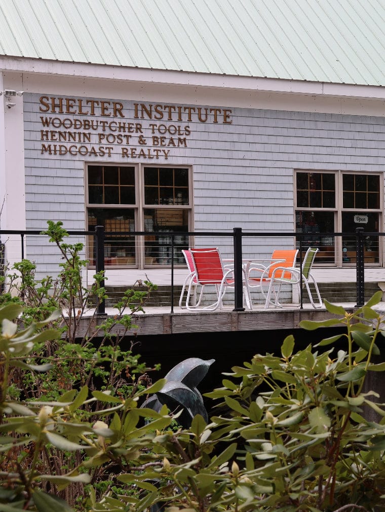 A patio with colorful chairs sits outside a gray building with signs for Shelter Institute, shelter tools, Henin Post & Beam, and Midcoast Realty. Green plants and a metal sculpture add charm to the scene.