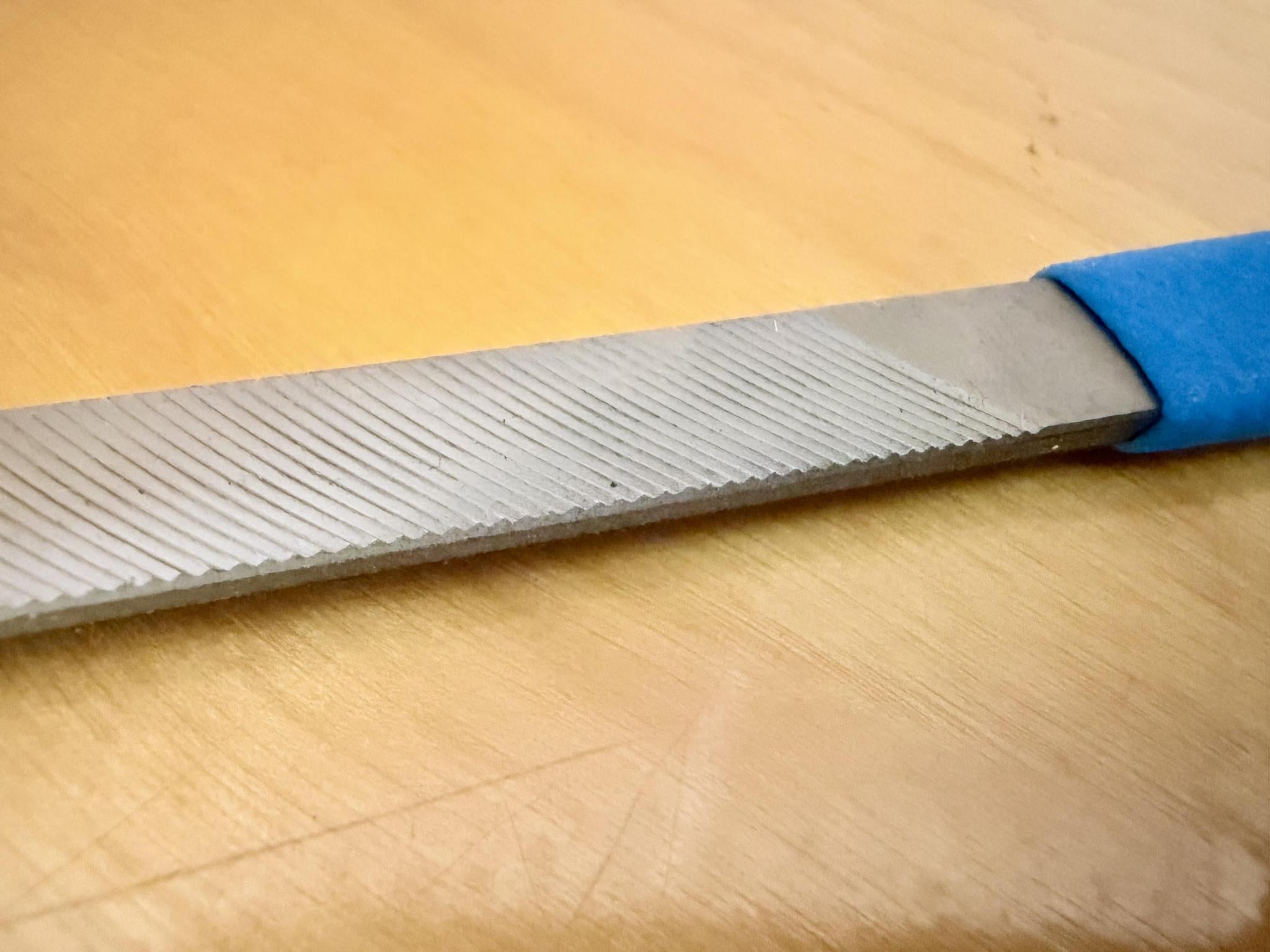 A close-up of the Iwasaki Draw File by Iwasaki, featuring diagonal ridges and a blue handle on light wood—a must-have tool for precise wood carving and cabinet making.