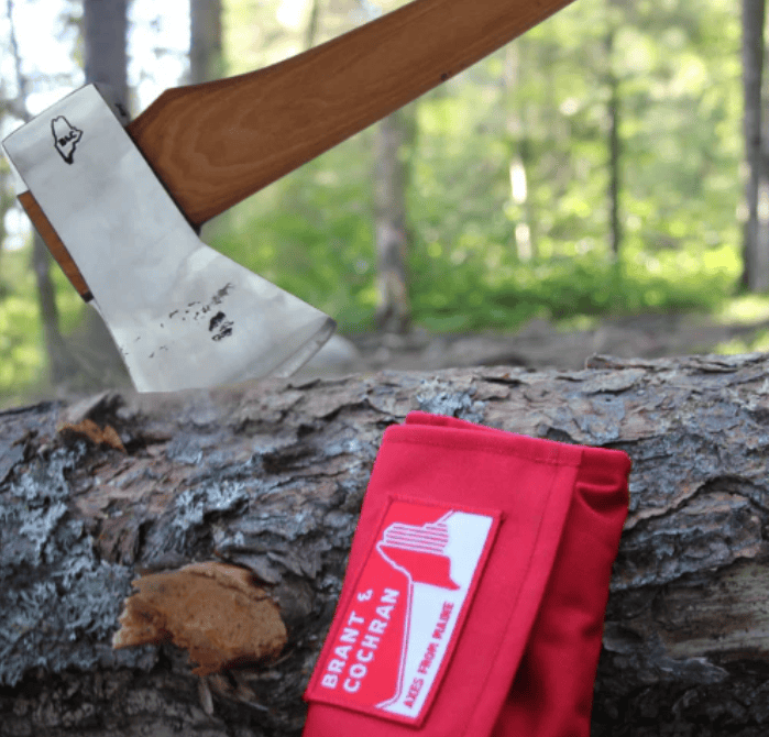A close-up of The Voyageur axe by BNC, embedded in a log with its hickory handle visible and a red blade cover nearby, set against a sunlit, green forest backdrop.