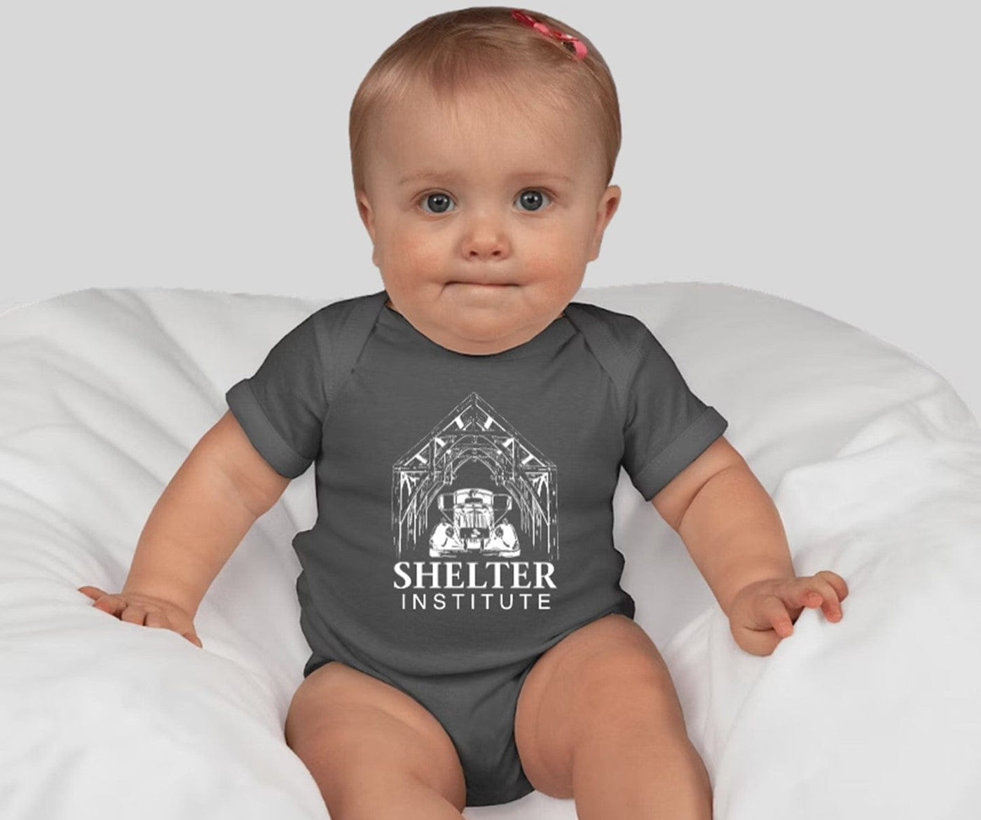 A baby with light hair and a small pink bow sits on a white cushion, wearing a dark gray Custom Ink Shelter Onesie featuring the Shelter Institute name and house graphic on the front.