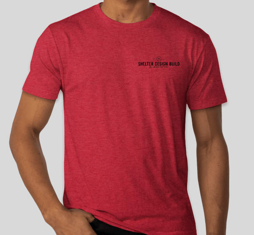 A person wears a red Custom Ink Shelter Mack Truck T-Shirt with small black text and a circular logo, standing against a plain light gray background that emphasizes its Hammer Beam Truss Frame inspiration from Woolwich, Maine.