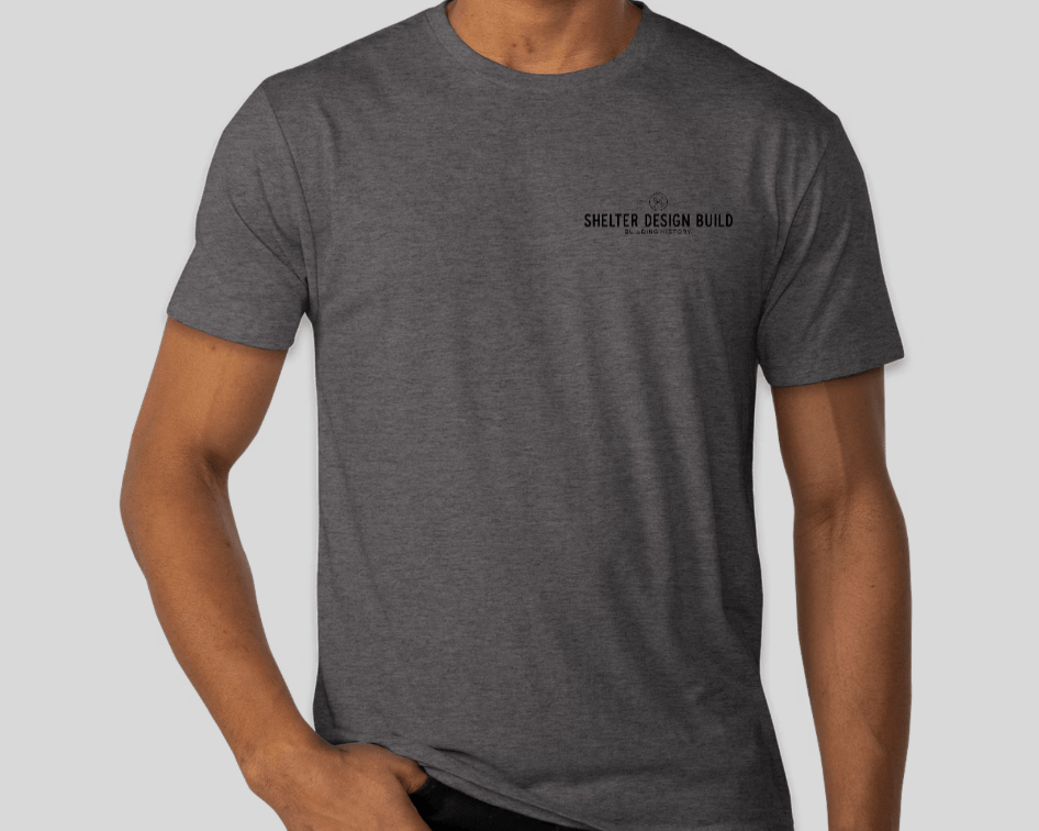A person wears a Custom Ink Shelter Mack Truck T-Shirt, plain dark gray with Shelter Design Build and logo on the upper left chest. The light gray background is inspired by Hammer Beam Truss Frame designs from Woolwich, Maine.