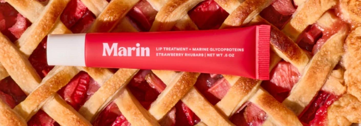 A red Marin Lip Treatment tube by Marin sits atop a golden strawberry rhubarb pie, hinting at the hydrating balm inside enriched with marine glycoproteins.
