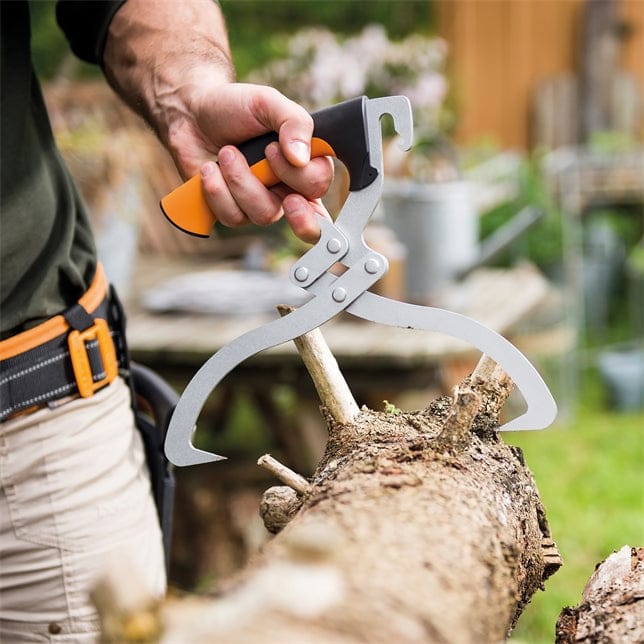 A person uses Fiskars Log Tongs by FIS, featuring an orange and black handle and made from hardened boron steel, to grip and lift a tree branch outdoors while wearing a tool belt in a garden.
