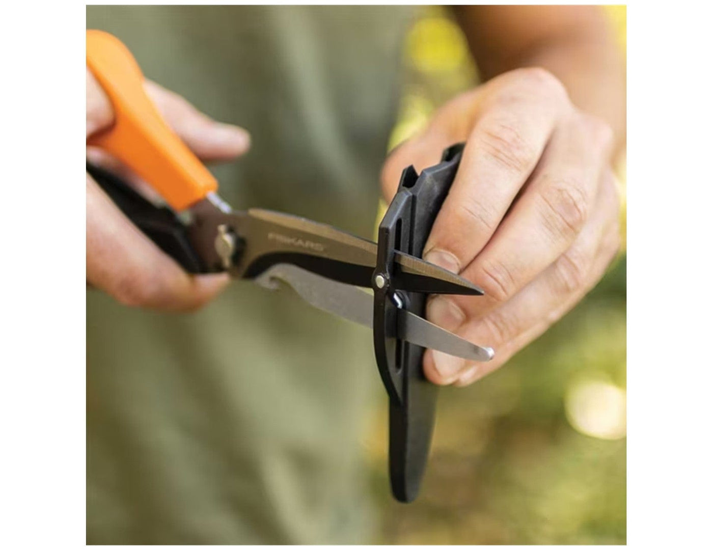 A person uses FIS Multi Purpose Garden Shears with precision-ground steel blades and orange handles to cut black plastic packaging outdoors, with a blurred green background.