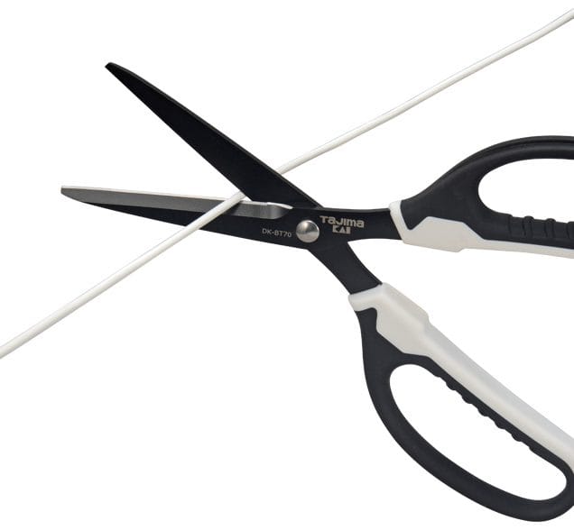 A pair of Fiskars Tajima VARIX Non-Stick Scissors with adhesive-resistant blades is shown cutting a white cable against a clean, white background.