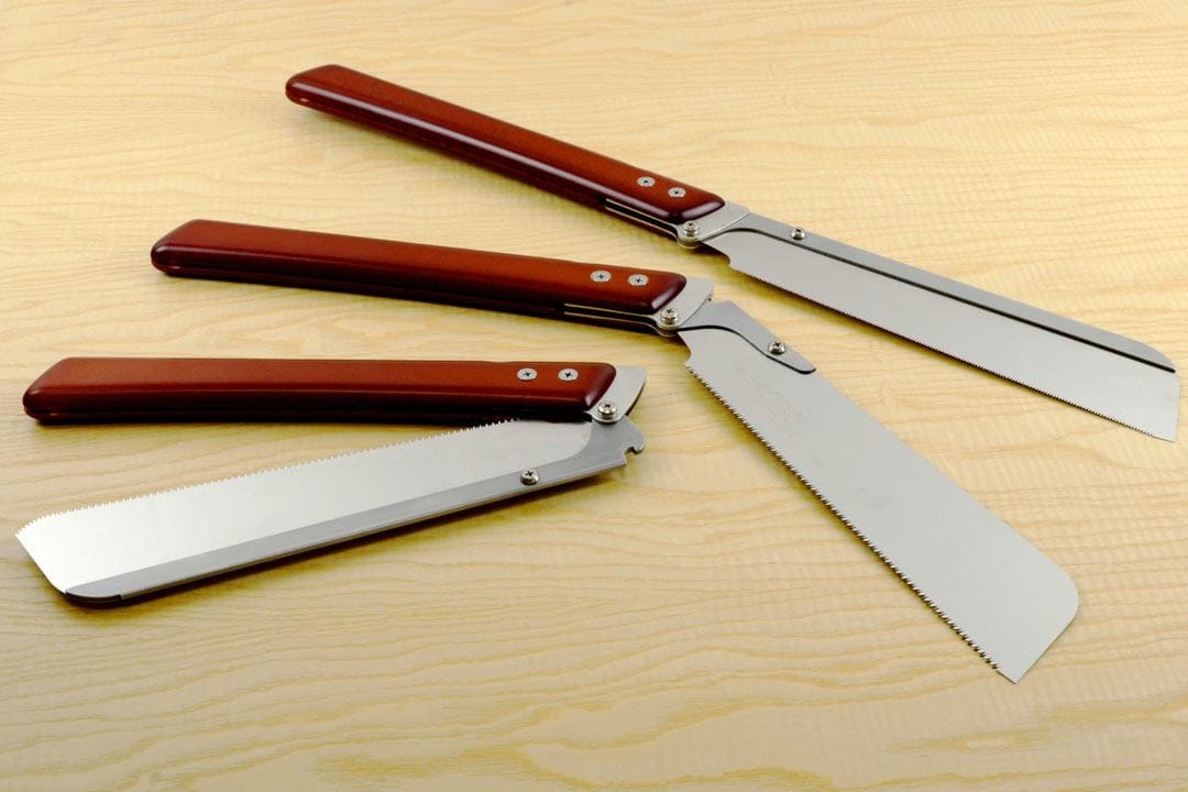 Three Shelter Institute Fugaku Folding Saws with red handles rest on a light wood surface; two are open, displaying straight and serrated blades, and one is closed with only its handle visible.