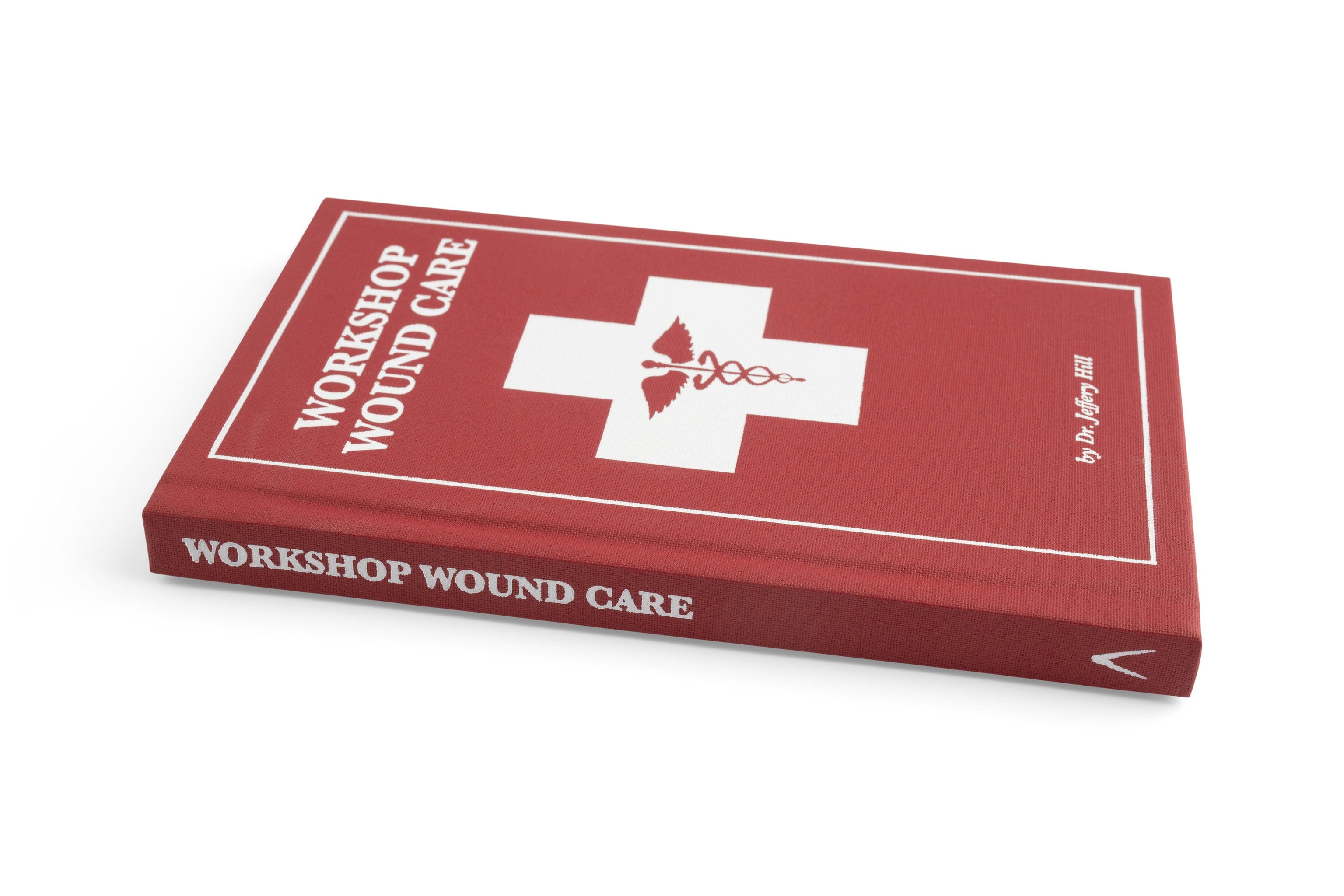 A red hardcover book titled Workshop Wound Care by LAP, focused on first aid for woodworkers and featuring a white medical cross and caduceus symbol, lies on a white surface. The authors name appears in white text on the cover.