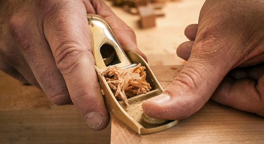 Close-up of hands using the Lie-Nielsen Low Angle Block Plane No 102 to shave wood curls from a board, showcasing Lie-Nielsen’s precision craftsmanship in woodworking.