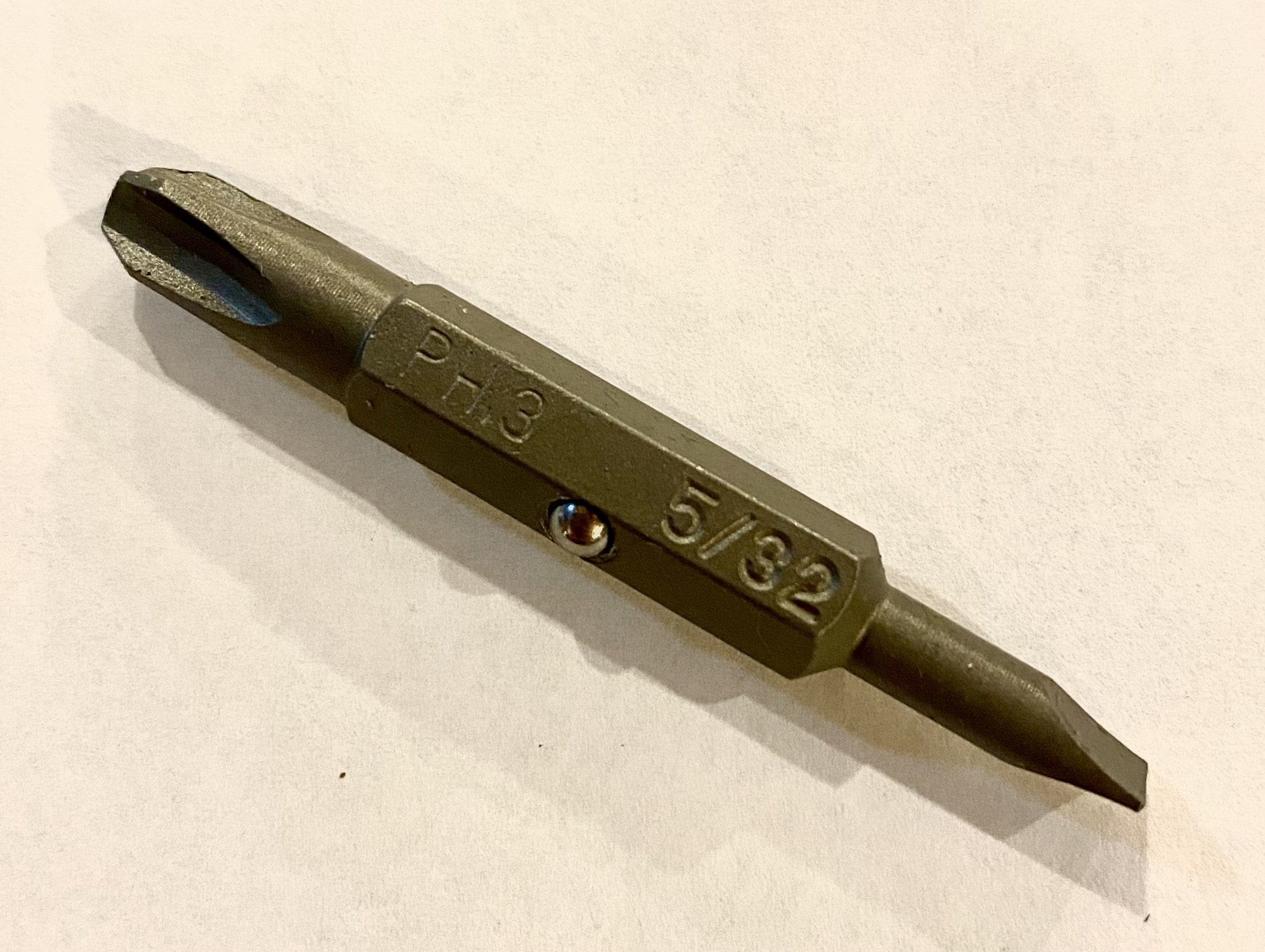 A LUT Shelter Tools Lutz Screwdriver with a reversible bit, featuring one end as a Phillips PH3 and the other as a 5/32 slotted head, lies on a white surface.