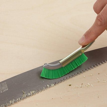 A hand uses the MARU Japanese File Cleaning Brush with nylon bristles to remove sawdust from a saw blade on a light wooden surface.