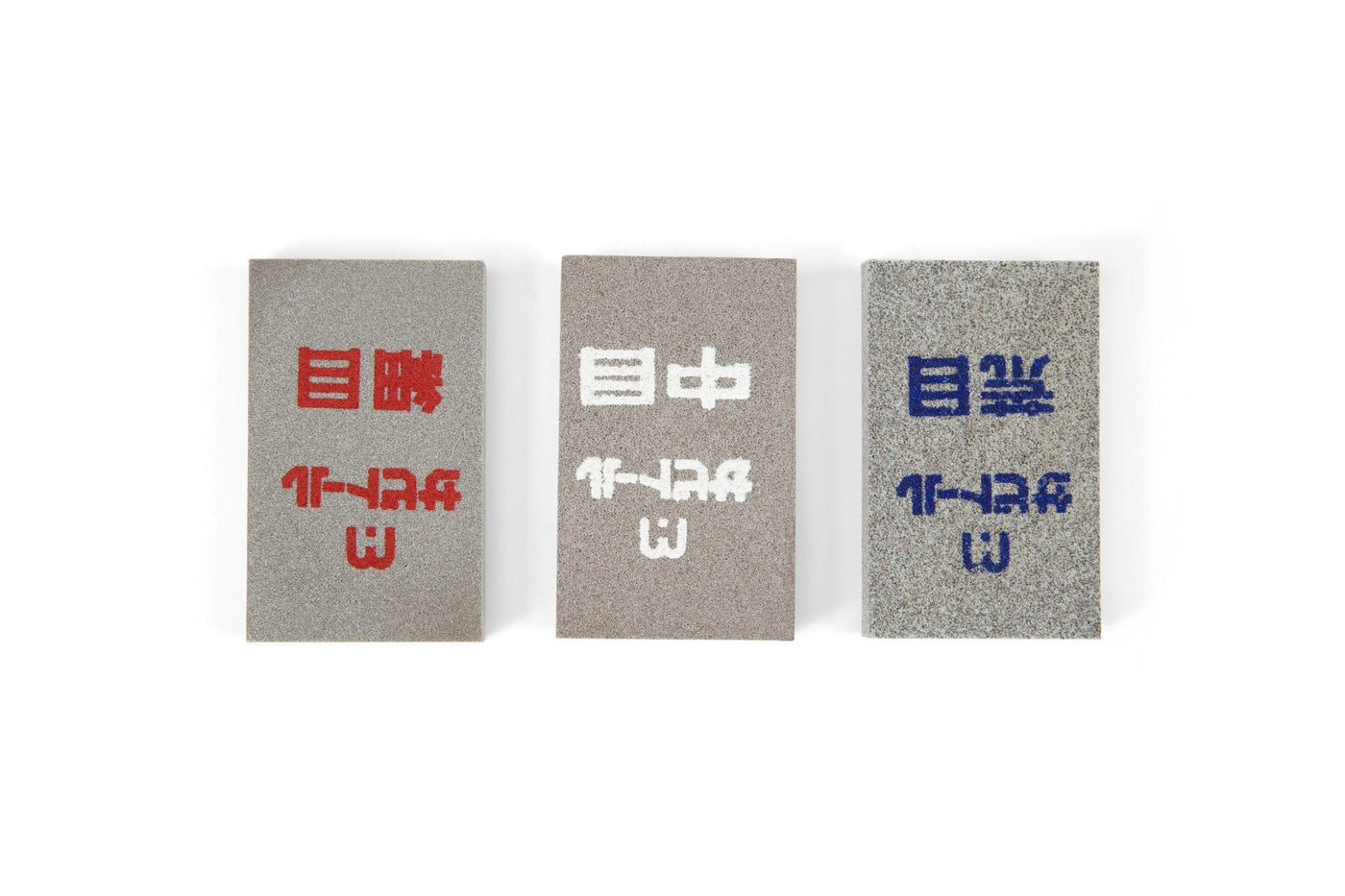 Three rectangular MARU Japanese Rust Erasers with rough textures are arranged side by side. Each stone, for rust removal on steel tools, displays Japanese kanji and markings in red, white, or blue. The background is white.