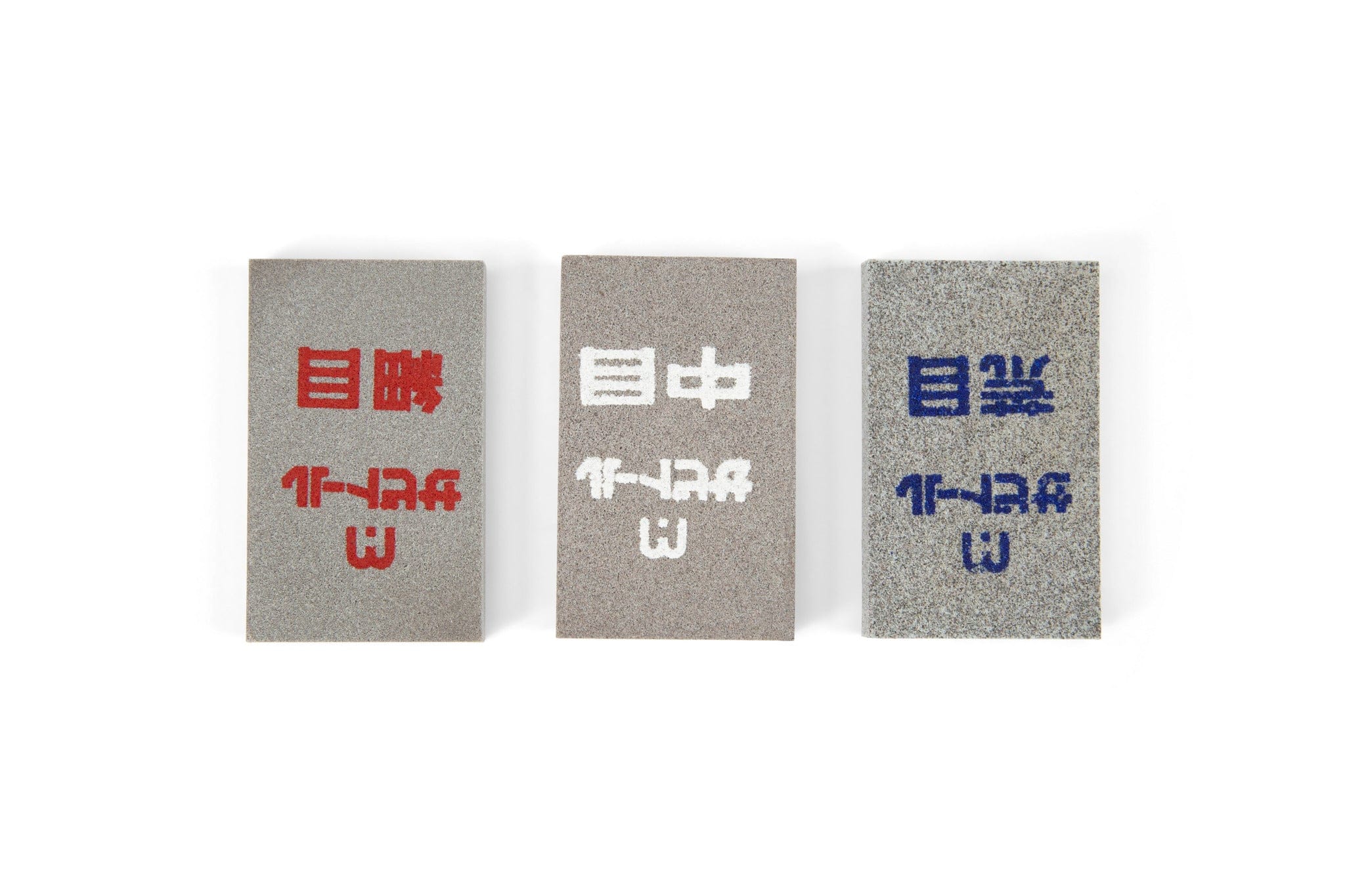Three rectangular MARU Japanese Rust Erasers with rough textures are arranged side by side. Each stone, for rust removal on steel tools, displays Japanese kanji and markings in red, white, or blue. The background is white.
