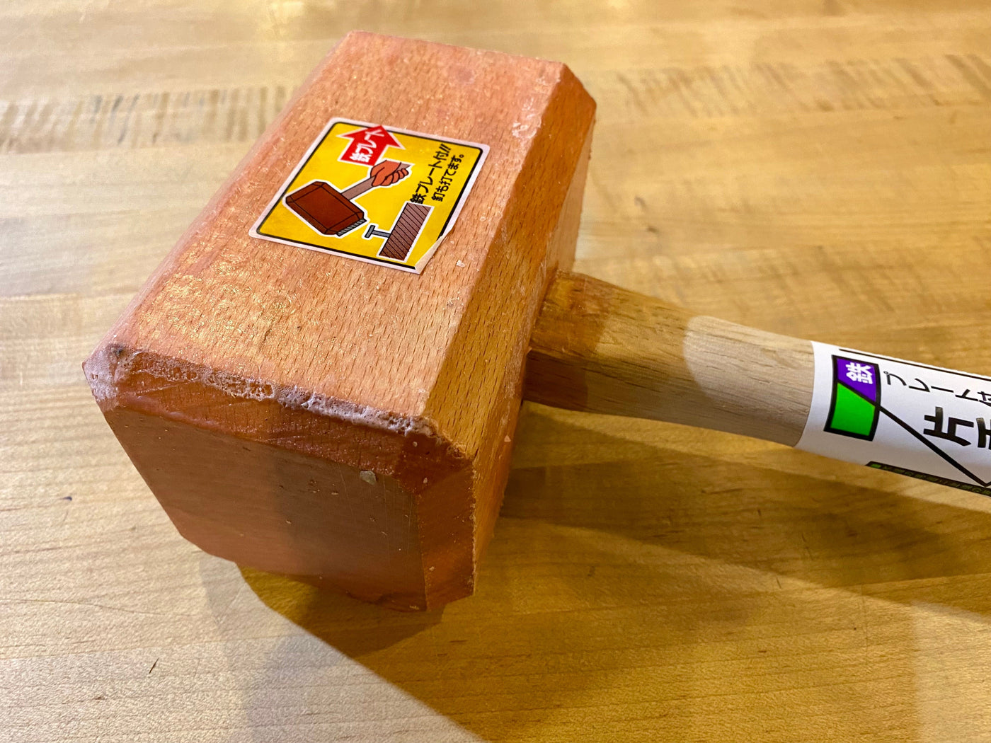 The MARU Iron-Plated Wood Mallet, made from Japanese White Oak, features a rectangular head with a handle and an illustrated sticker in Japanese. It rests elegantly on a wooden surface.