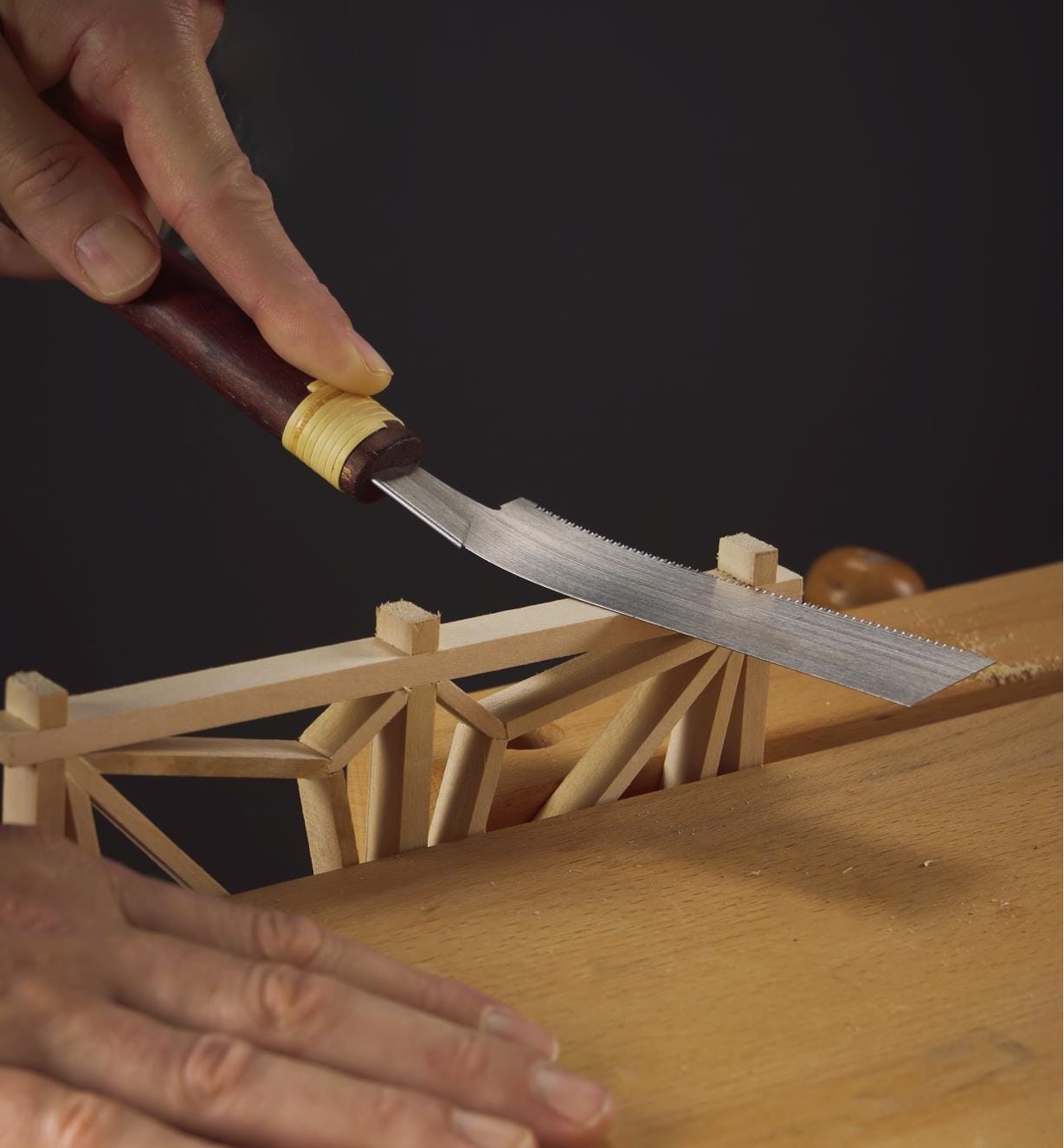 A hand skillfully uses the MARU Kugihiki Flush Saw to precisely cut a small wooden structure on the workbench.