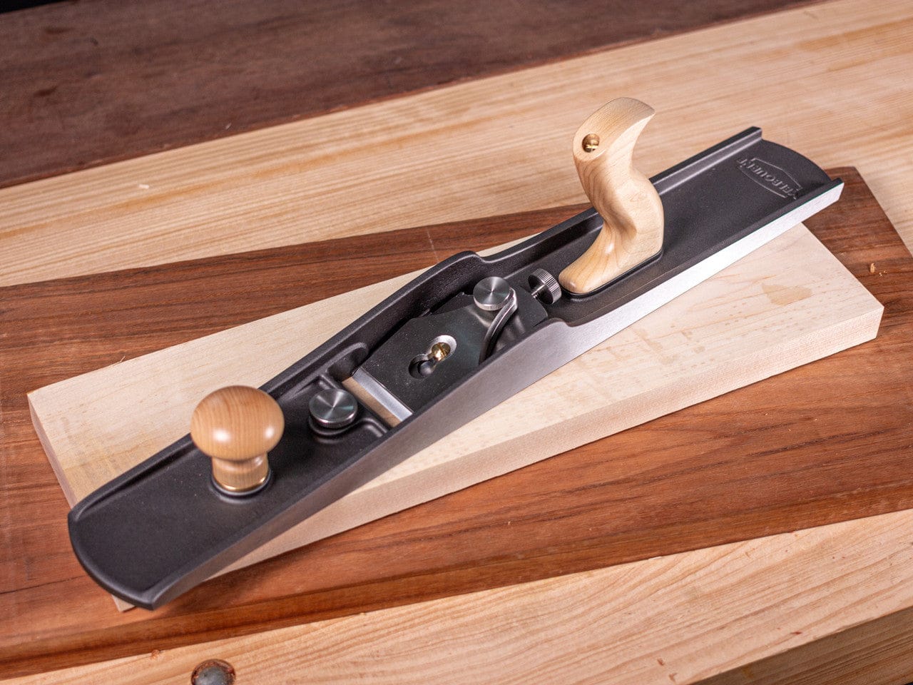 A Melbourne Tool Company Low Angle Jointing Plane with wooden handles sits on a workbench, showcasing fine woodworking craftsmanship.
