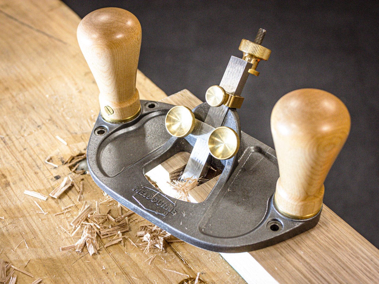 A Melbourne Tool Router Plane by the Melbourne Tool Company is depicted smoothing a wooden surface, with wood shavings around it, featuring wooden handles.