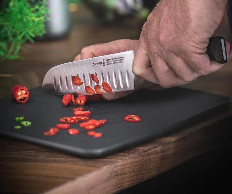 A hand skillfully using an Opinel Le Forges 1890 Kitchen Knife effortlessly slices red chili peppers on a black cutting board, showcasing precision and elegance.