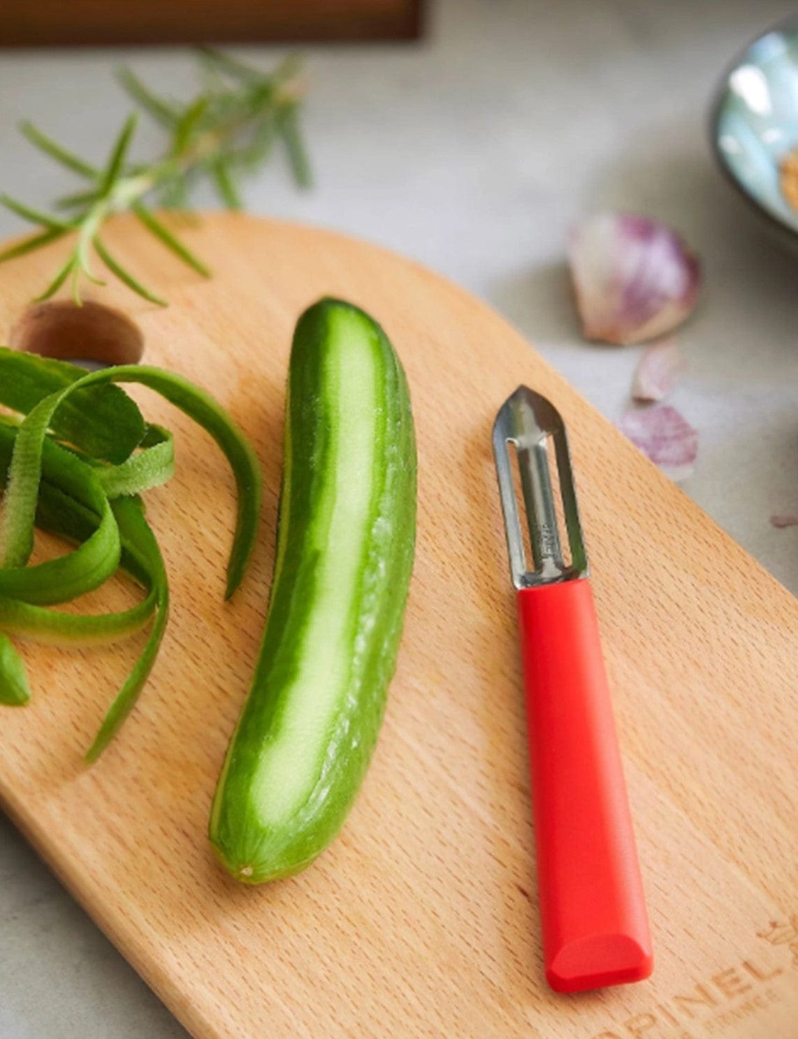 A half-peeled cucumber alongside an Opinel Essential Stationary Peeler with a stainless steel blade rests on a wooden cutting board, surrounded by cucumber peels and garlic, showcasing essential kitchen tools.