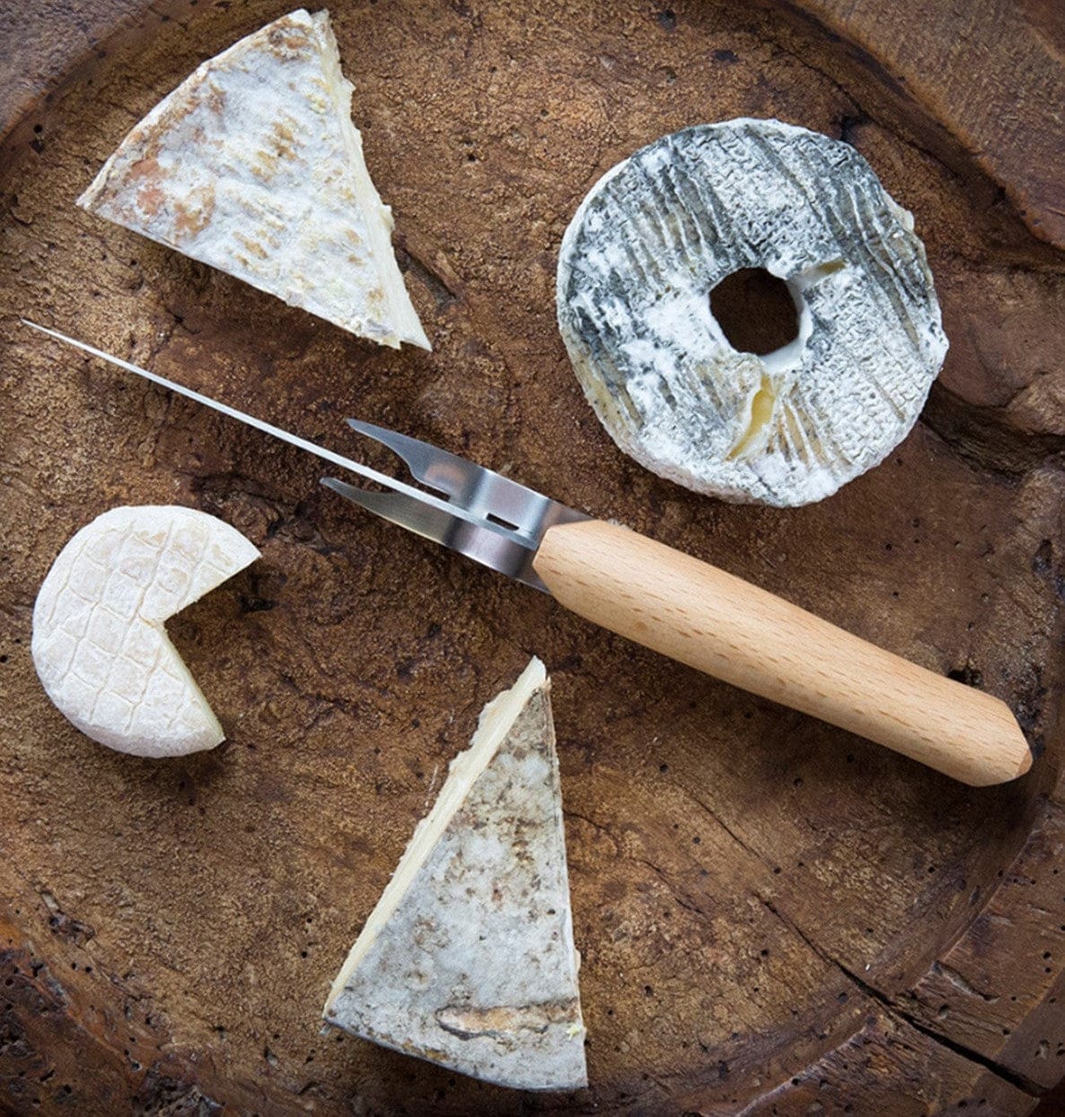 The Opinel Cheese Knife and Fork Set features four assorted cheeses with a stainless steel knife and beech wood handle, elegantly arranged on a wooden platter for a rustic presentation.