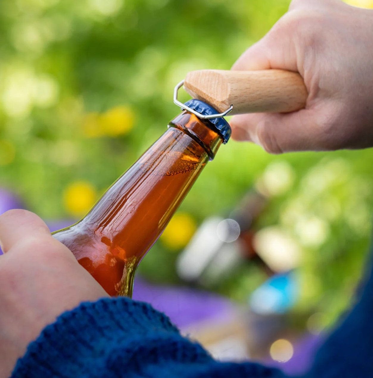 A person uses the Opinel No.10 Corkscrew Stainless Steel Folding Knife with Bottle Opener to open a brown glass bottle outdoors, with a green blurred background.