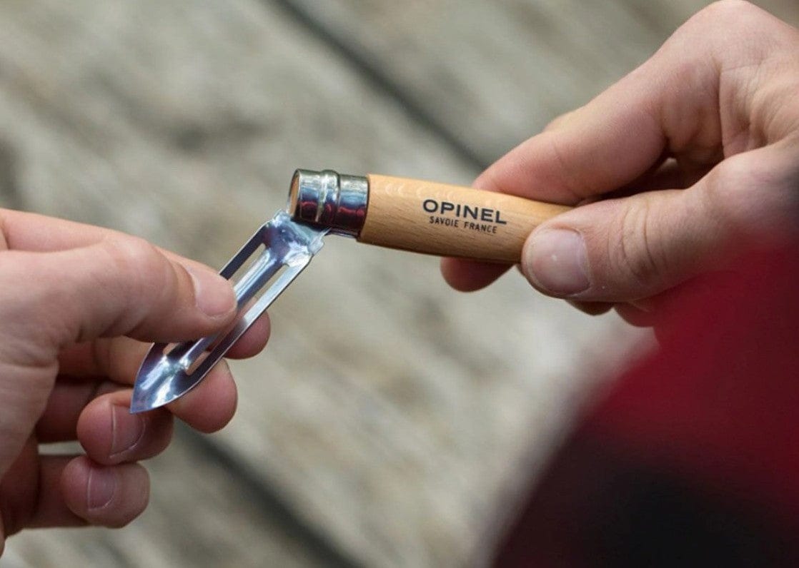 A person uses the Opinel Folding Peeler, with its wooden handle and stainless steel notched blade, to clean under their fingernail. Ideal for camp kitchen tasks or outdoor cooking, the peeler is branded “Opinel Savoie France.”.